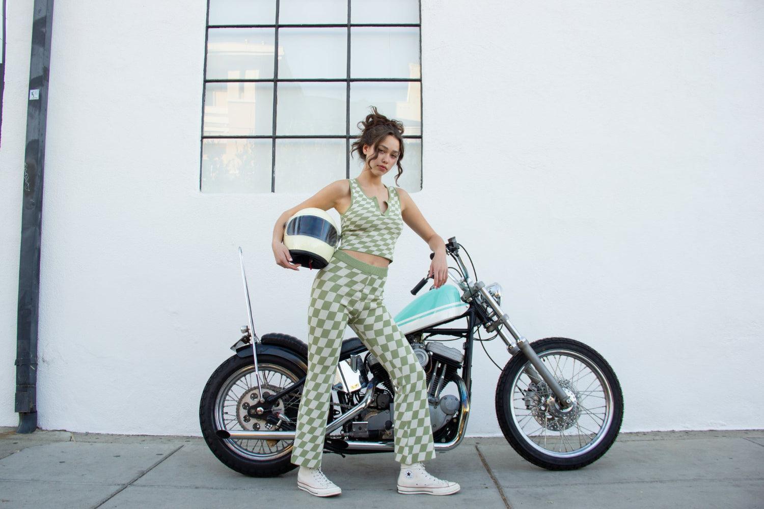 Young woman in green checkered outfit holding a white helmet, standing next to a vintage turquoise motorcycle.