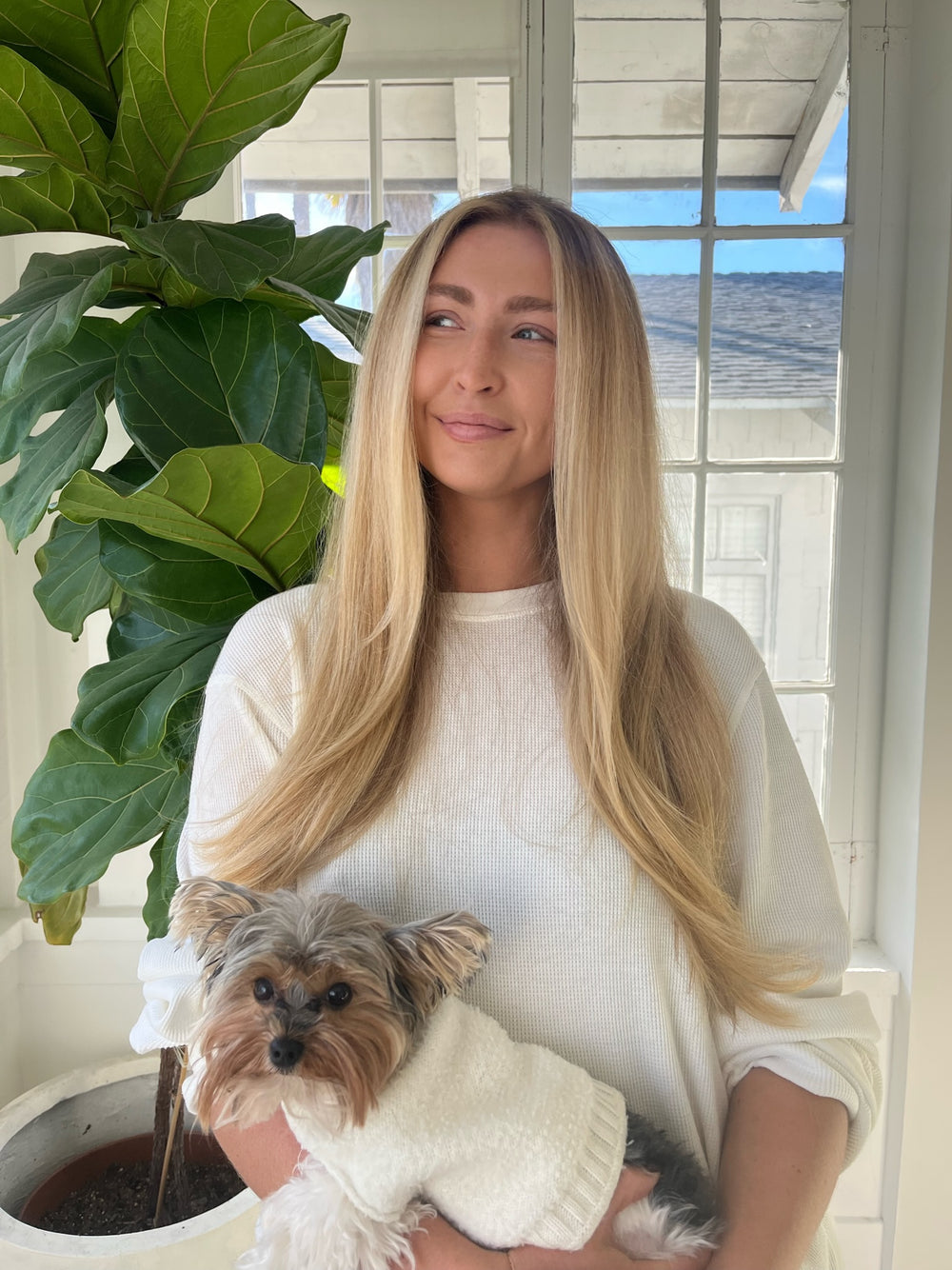 Blonde woman in a white waffle-knit top holding a small dog in a matching white sweater, looking left.
