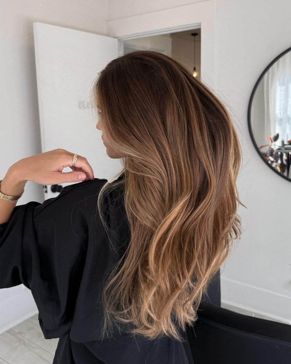 Woman with long, wavy brown hair featuring warm blonde balayage highlights, viewed from behind.