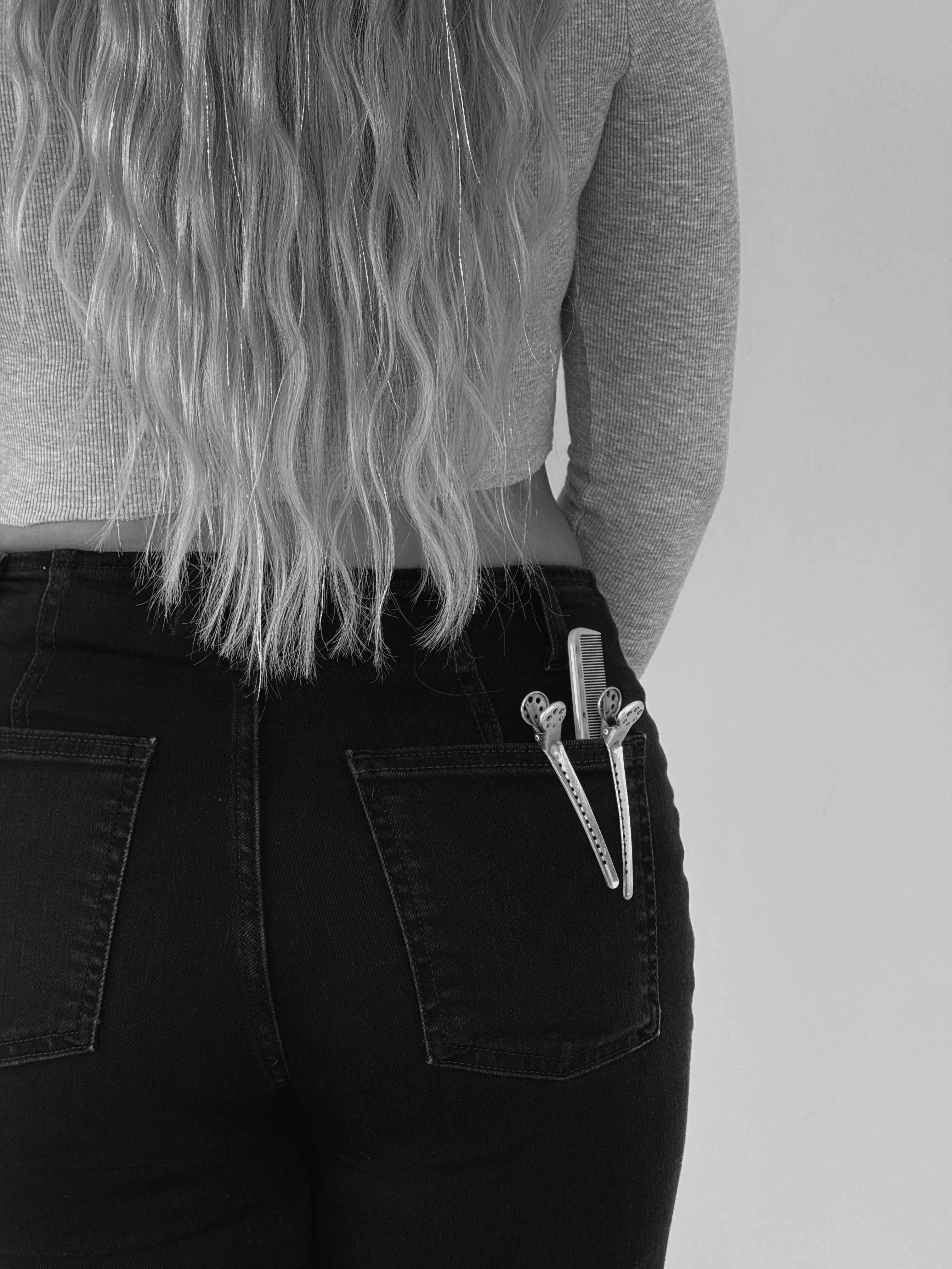 Black and white shot of a woman with long, wavy hair and tinsel, wearing black jeans with hair clips and a comb in her back pocket.