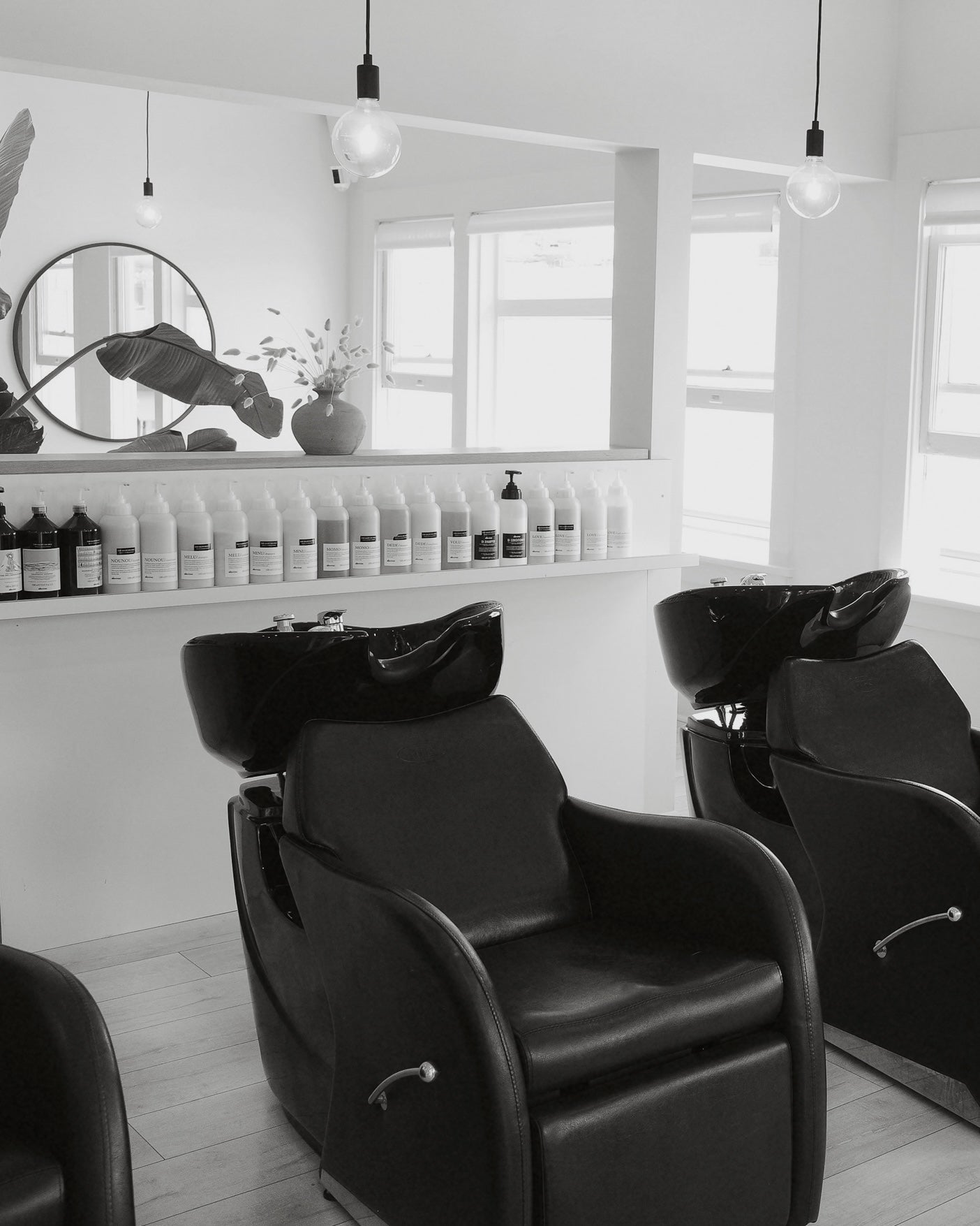 Black and white image of a modern hair salon with three shampoo stations, product display, and round mirror.