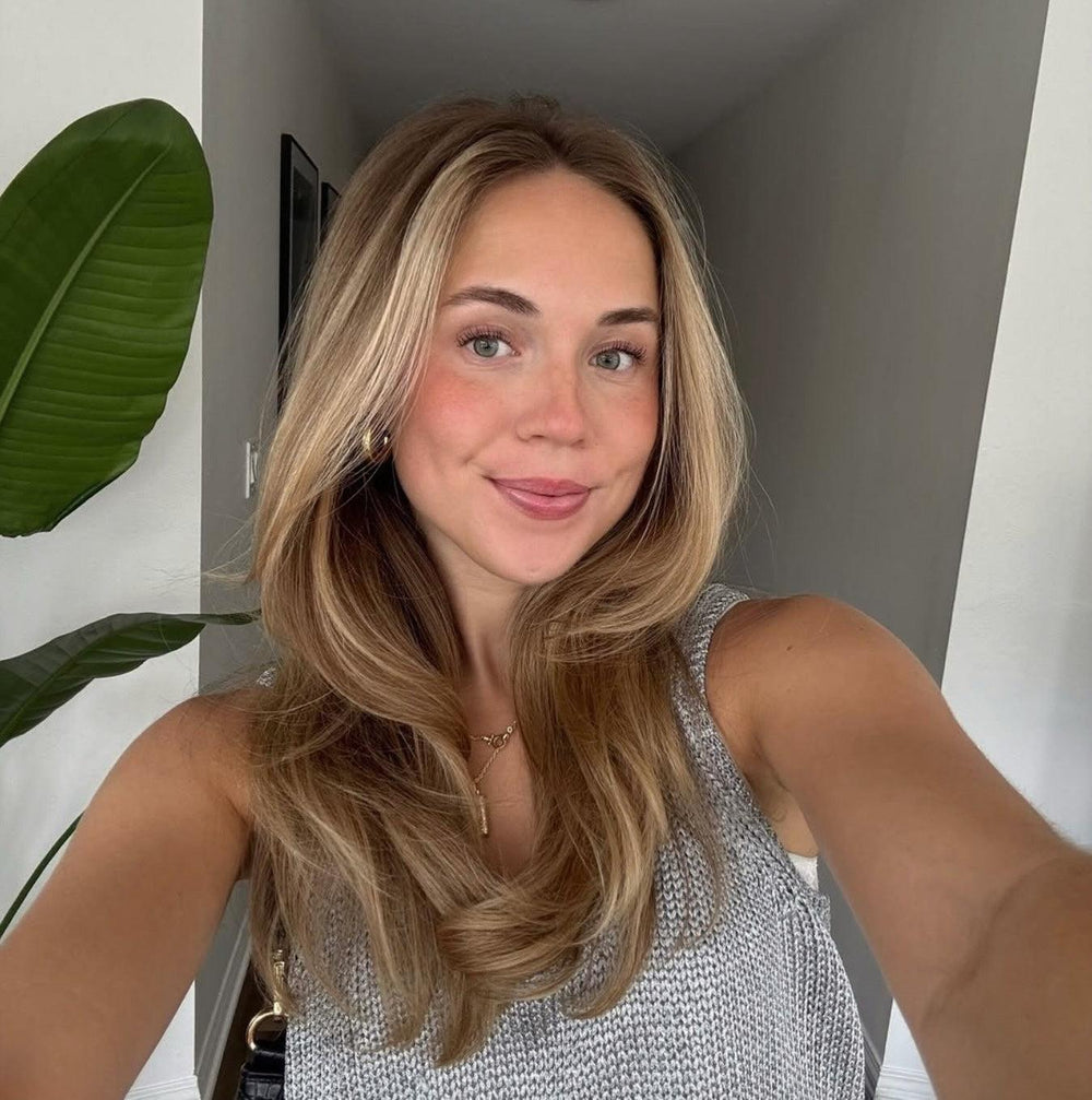 Smiling woman with long blonde hair and green eyes wearing a light gray knit tank top, next to a large green plant.