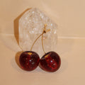 Gold cherry bomb earrings with deep red, glossy cherry charms, displayed against a clear quartz crystal.