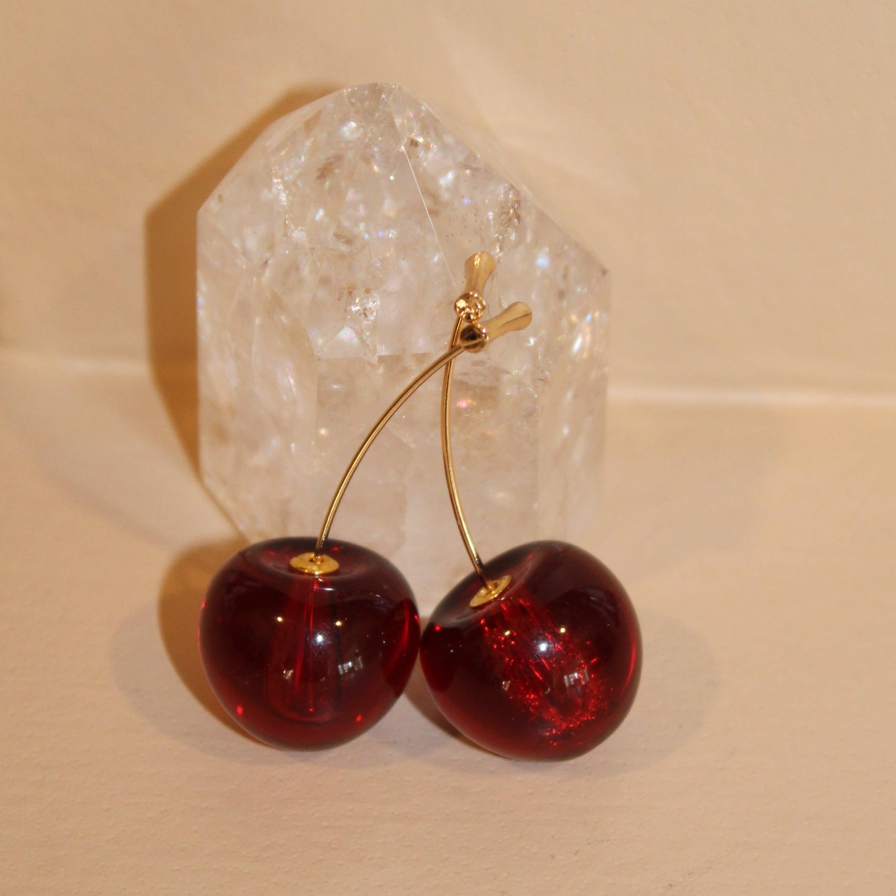Gold cherry bomb earrings with deep red, glossy cherry charms, displayed against a clear quartz crystal.