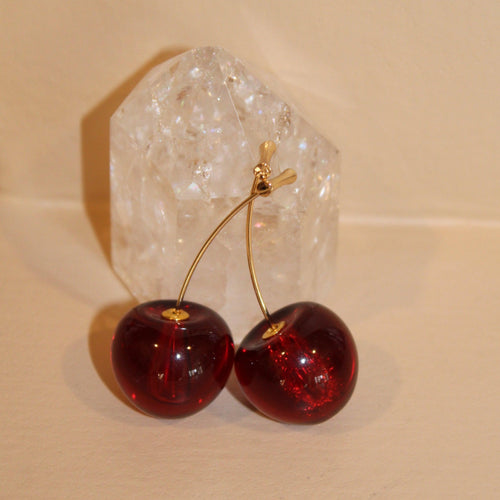 Gold cherry bomb earrings with deep red, glossy cherry charms, displayed against a clear quartz crystal.