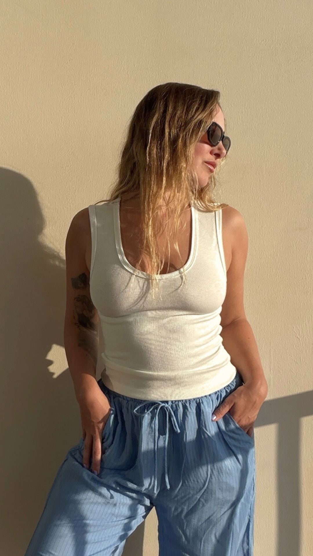 Woman in a white ribbed U-neck cotton Coco Tank top and blue pants, wearing sunglasses.