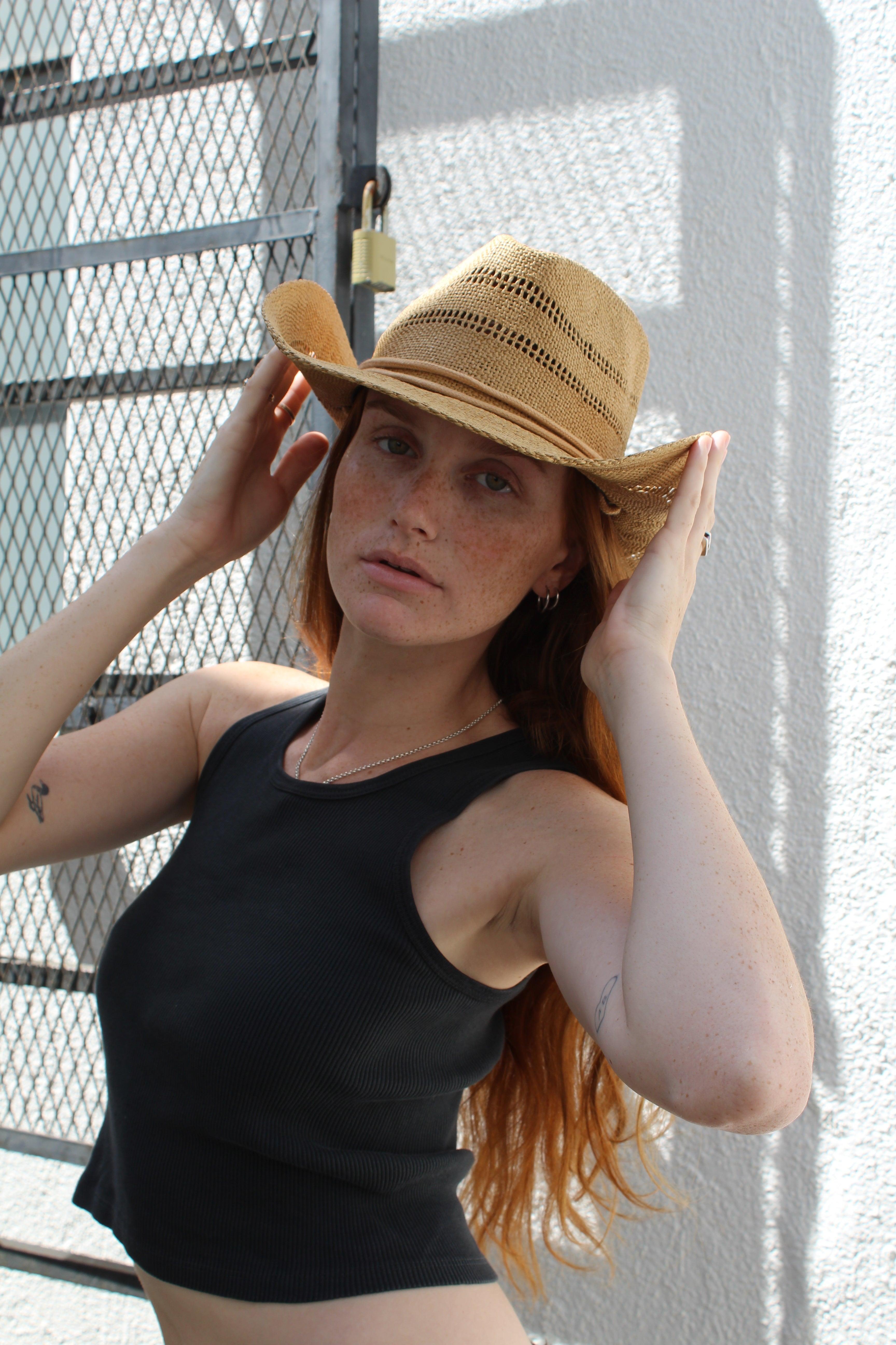 Woman with red hair wearing a light brown woven straw cowgirl hat and black tank top.