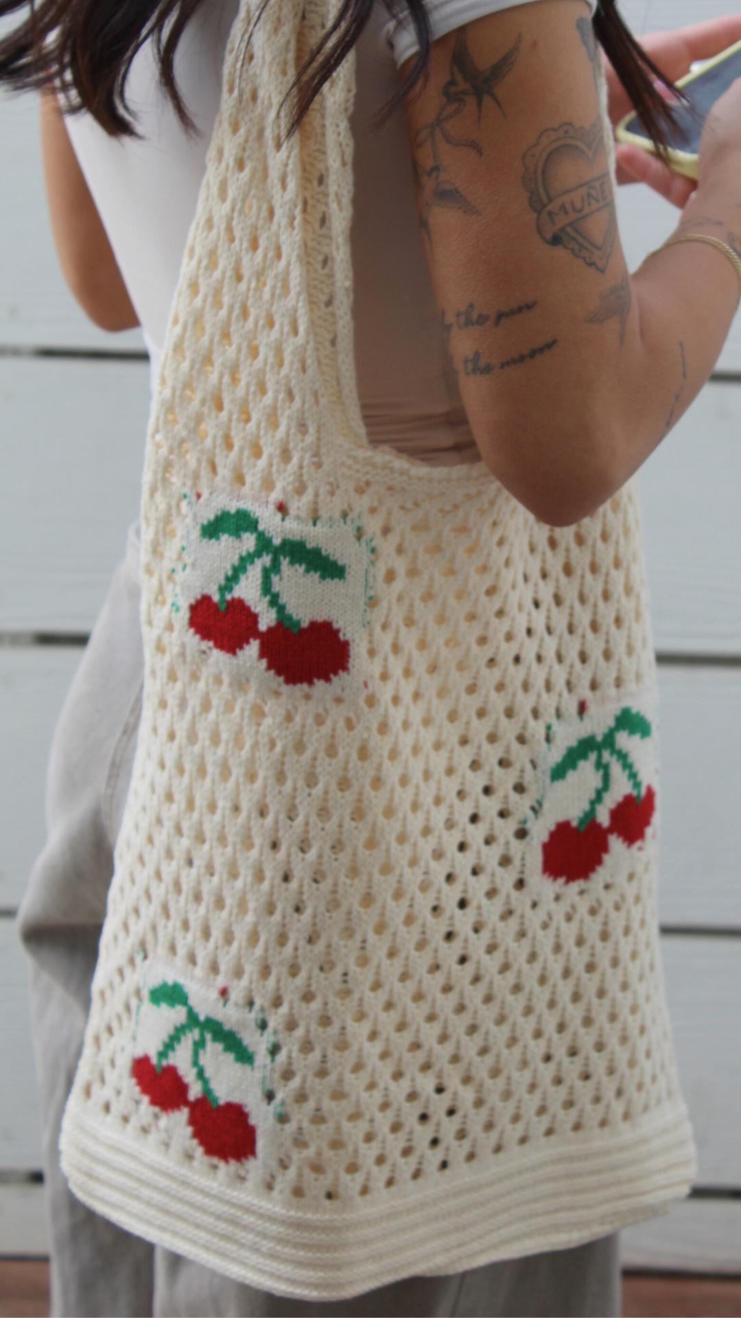 Cream crochet mesh fruit bag with red and green cherry designs, worn over a shoulder.
