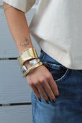 Thick, wavy golden and silver cuff bracelets stacked on a woman's arm, paired with jeans.