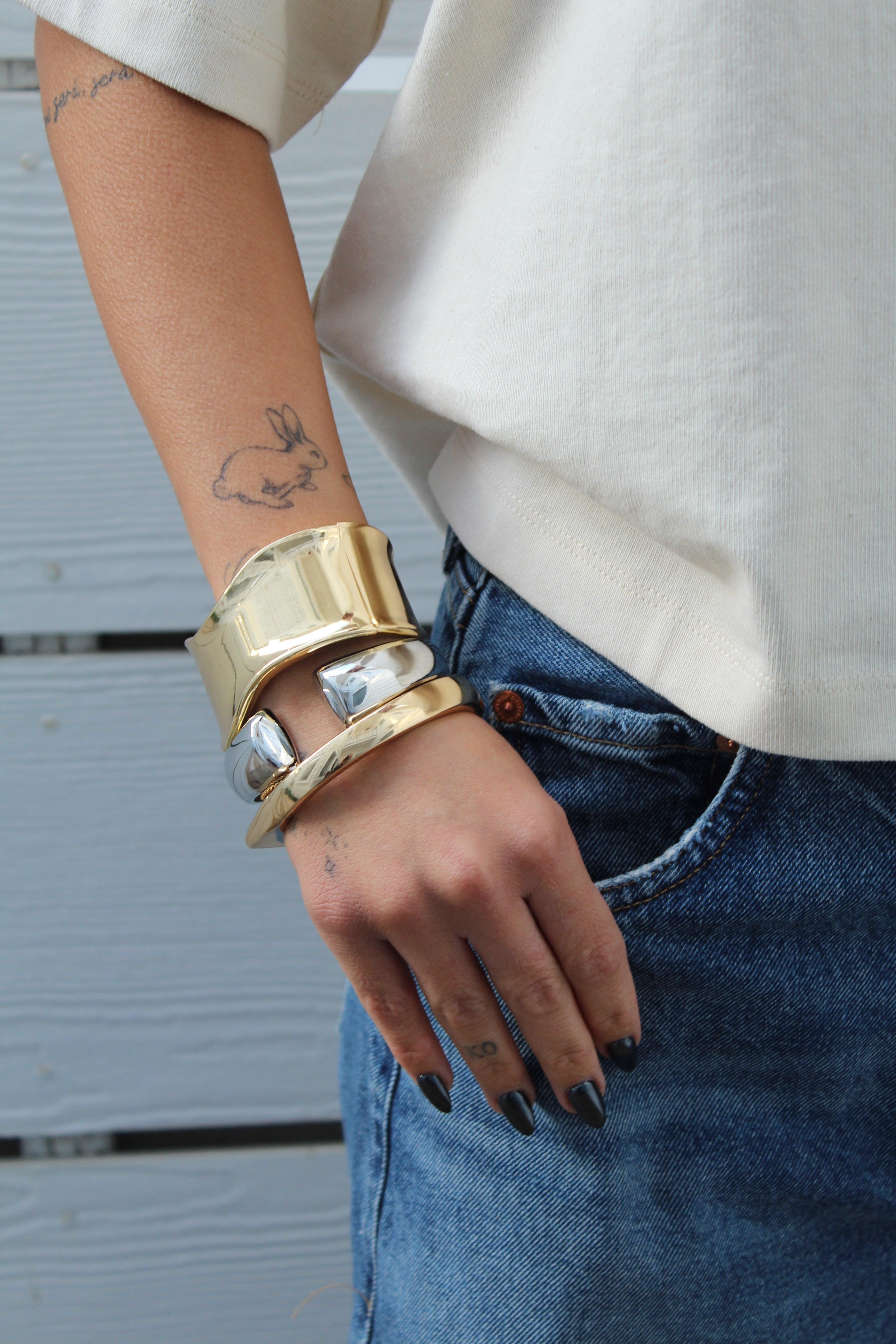 Thick, wavy golden and silver cuff bracelets stacked on a woman's arm, paired with jeans.
