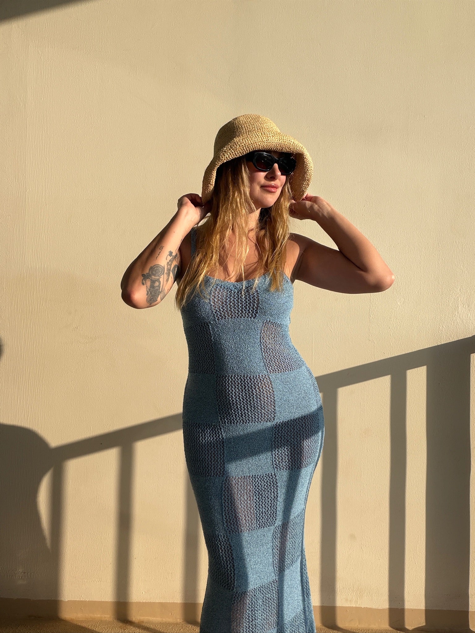 Model in blue knit maxi dress, straw hat, and sunglasses against a beige wall.