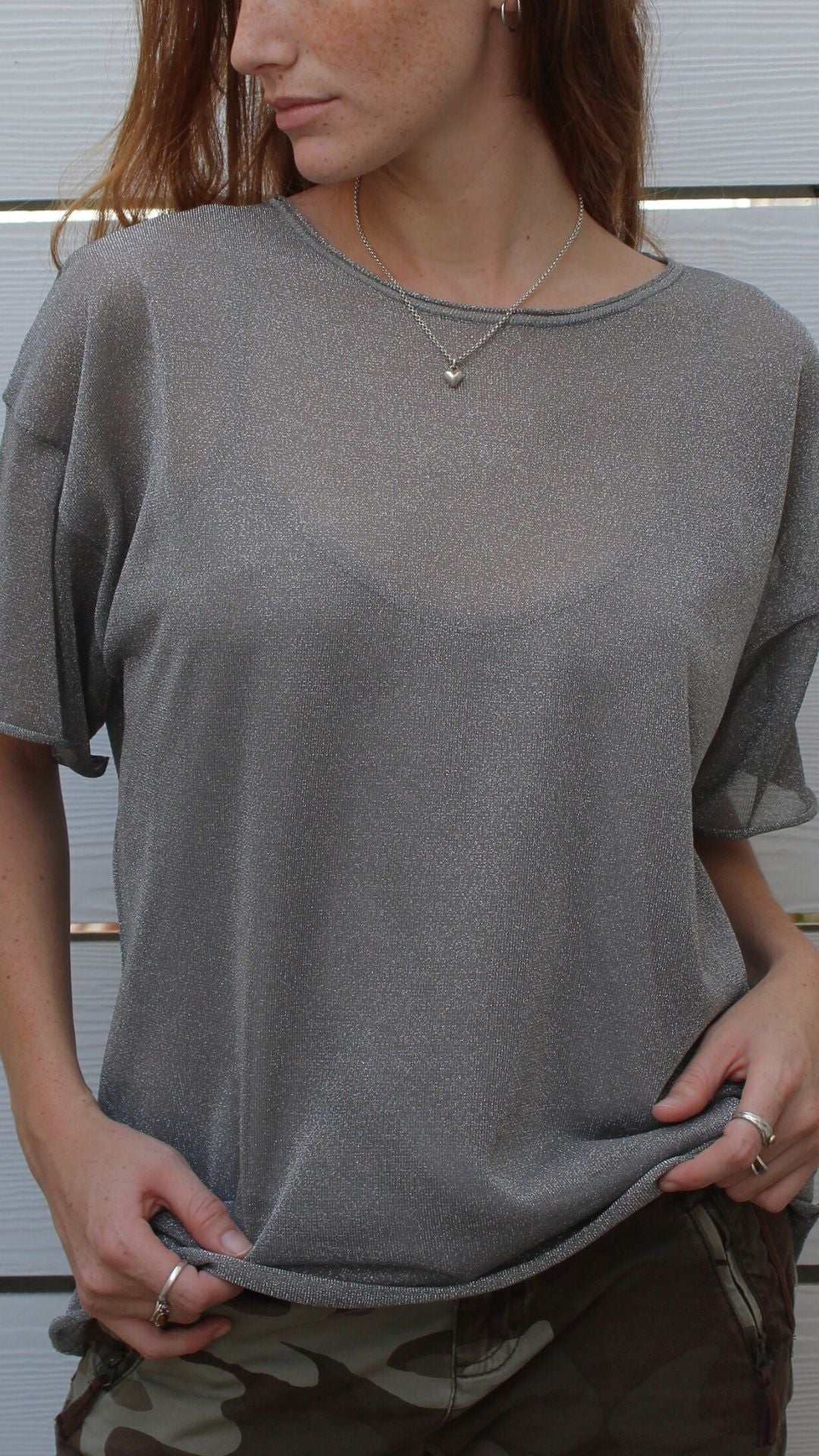 Woman in a sheer metallic silver boxy top with under tank, short sleeves, and a heart necklace.