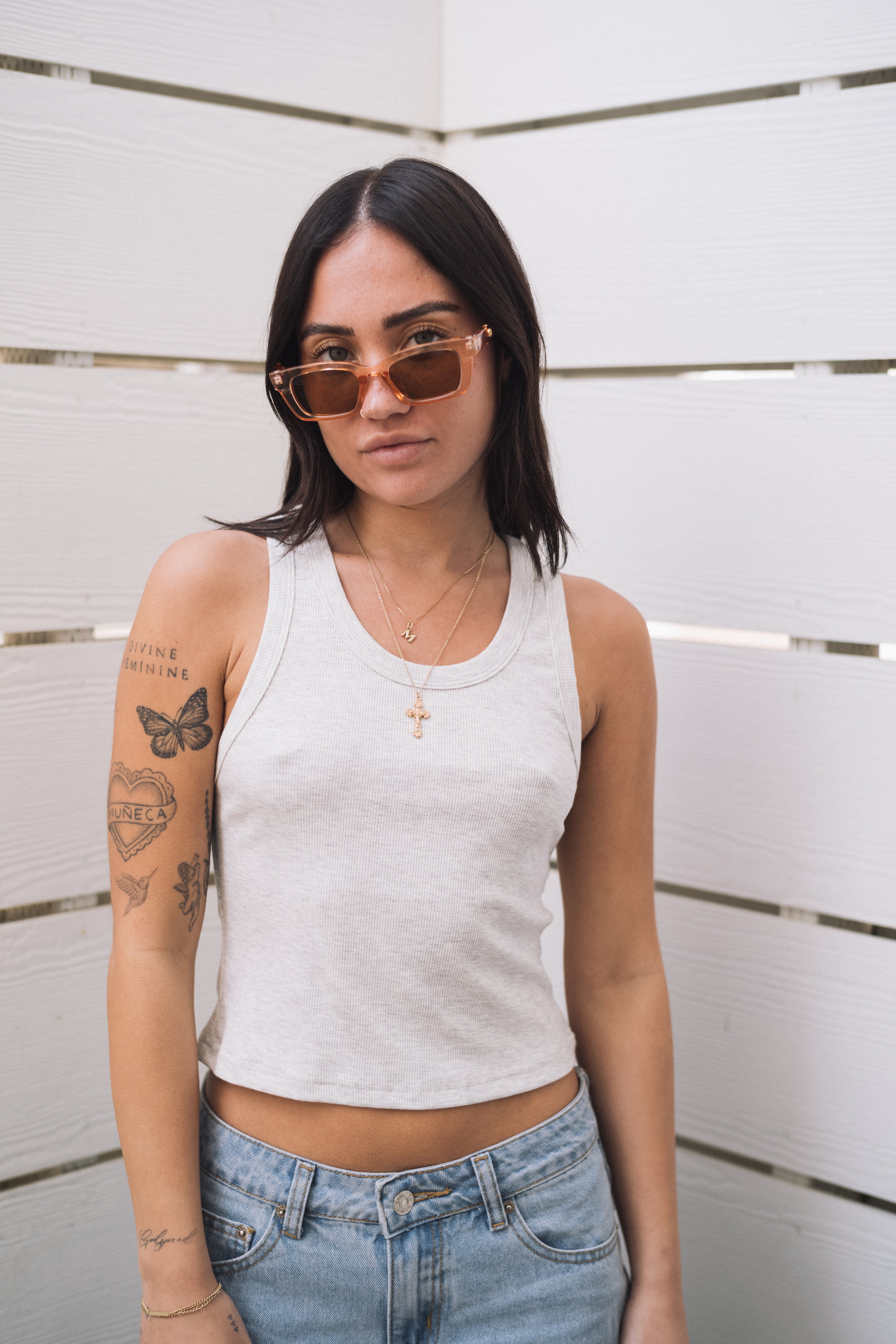 Woman in a ribbed fitted basic light grey tank top, light wash jeans, and orange sunglasses.