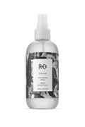 Clear R+Co Dallas Thickening Spray bottle, 8.5 oz, with a black and white label featuring text and a street scene.