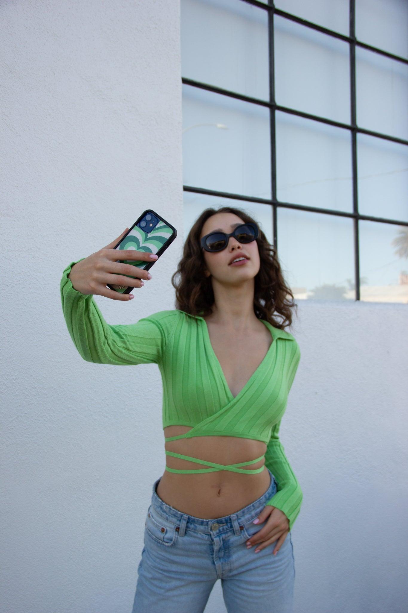 Model in a lime green ribbed front-tie long sleeve crop top, light wash jeans, and sunglasses, holding a phone.
