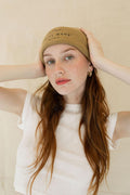 Woman with red hair wearing a tan Salty Beanie BB with black embroidery, white top, and gold earrings.