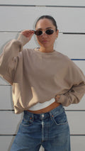 Model wearing a beige oversized crewneck sweatshirt, white top, and blue jeans, adjusting oval sunglasses.