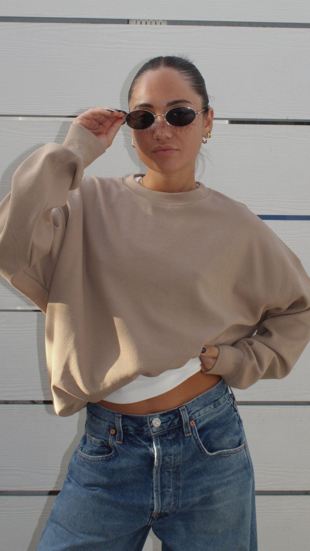 Model wearing a beige oversized crewneck sweatshirt, white top, and blue jeans, adjusting oval sunglasses.