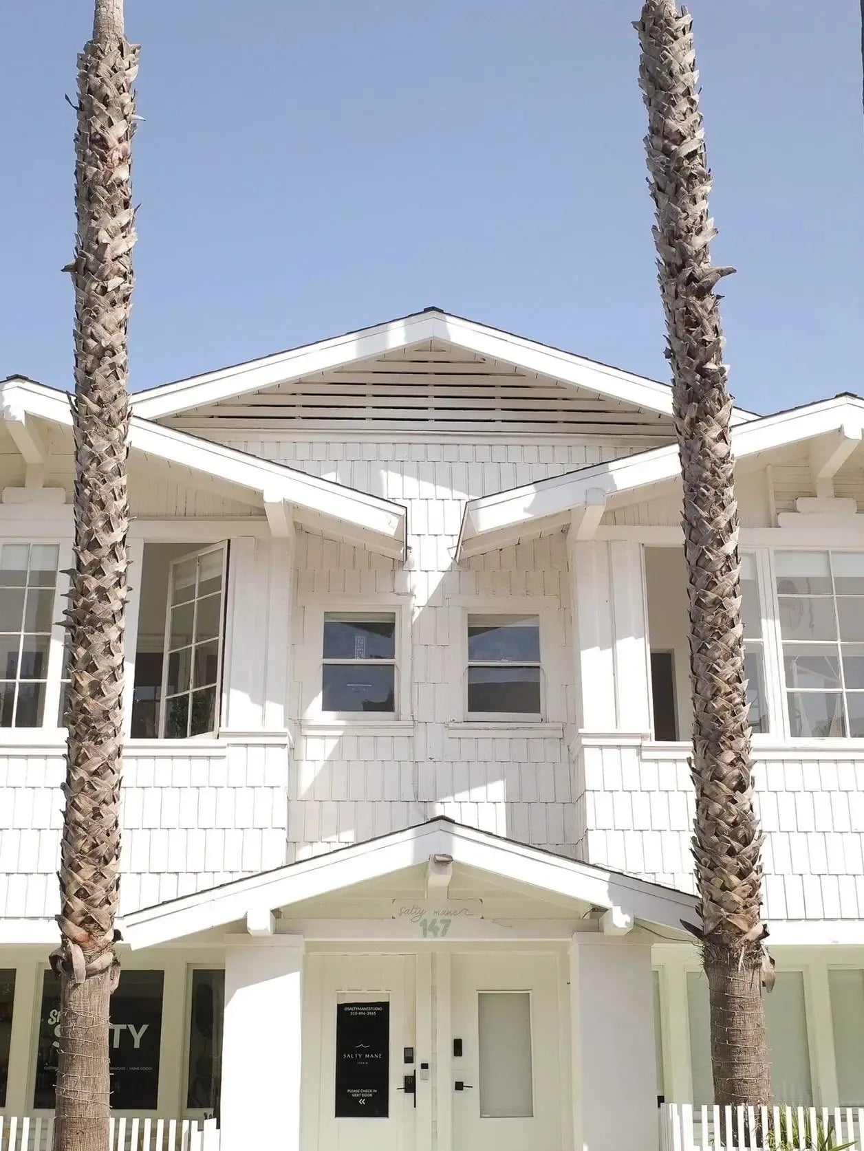 White shingle-sided beach house with multiple windows and a front door, framed by two tall palm tree trunks.