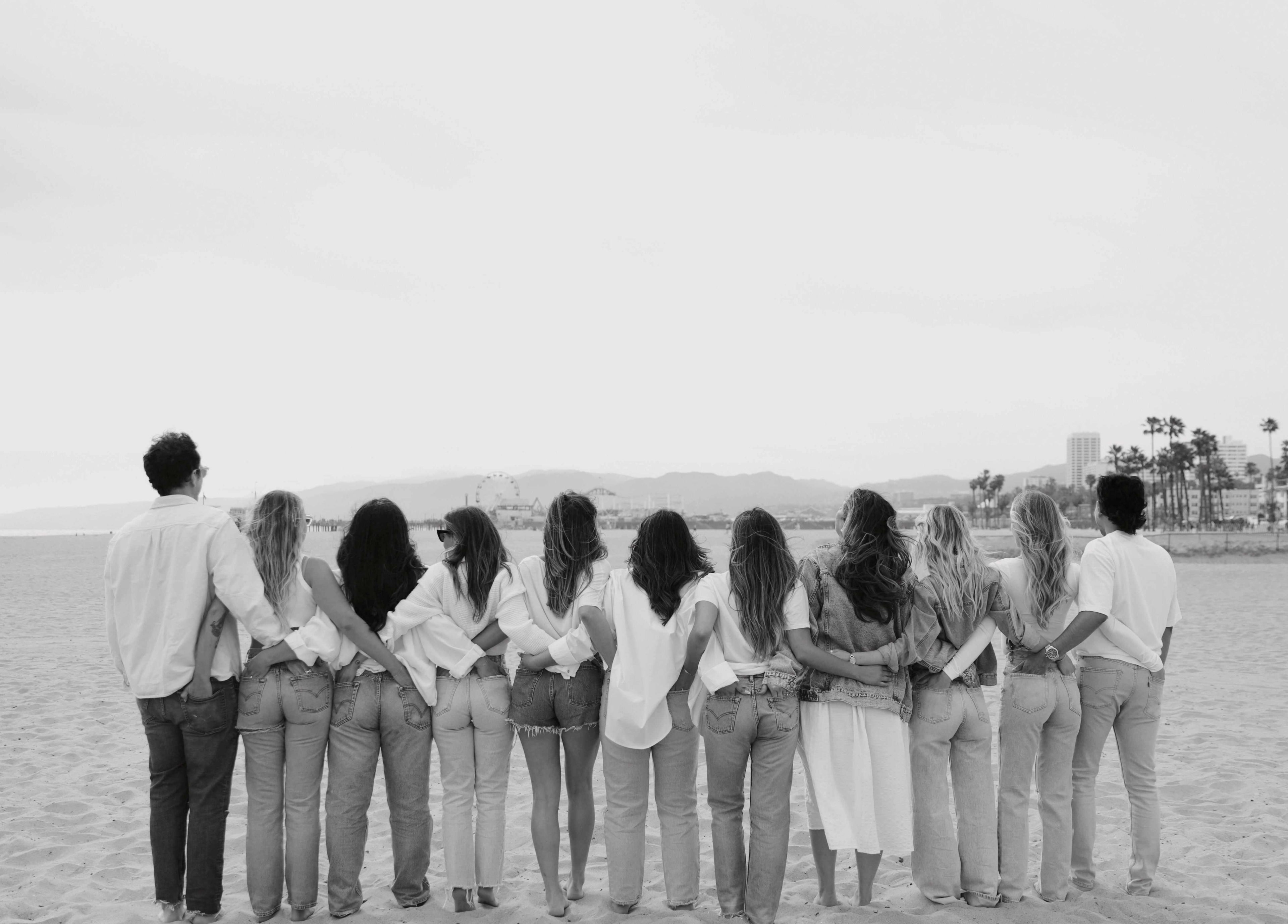 Ten people, mostly women, stand on a sandy beach with arms around each other, facing the ocean.