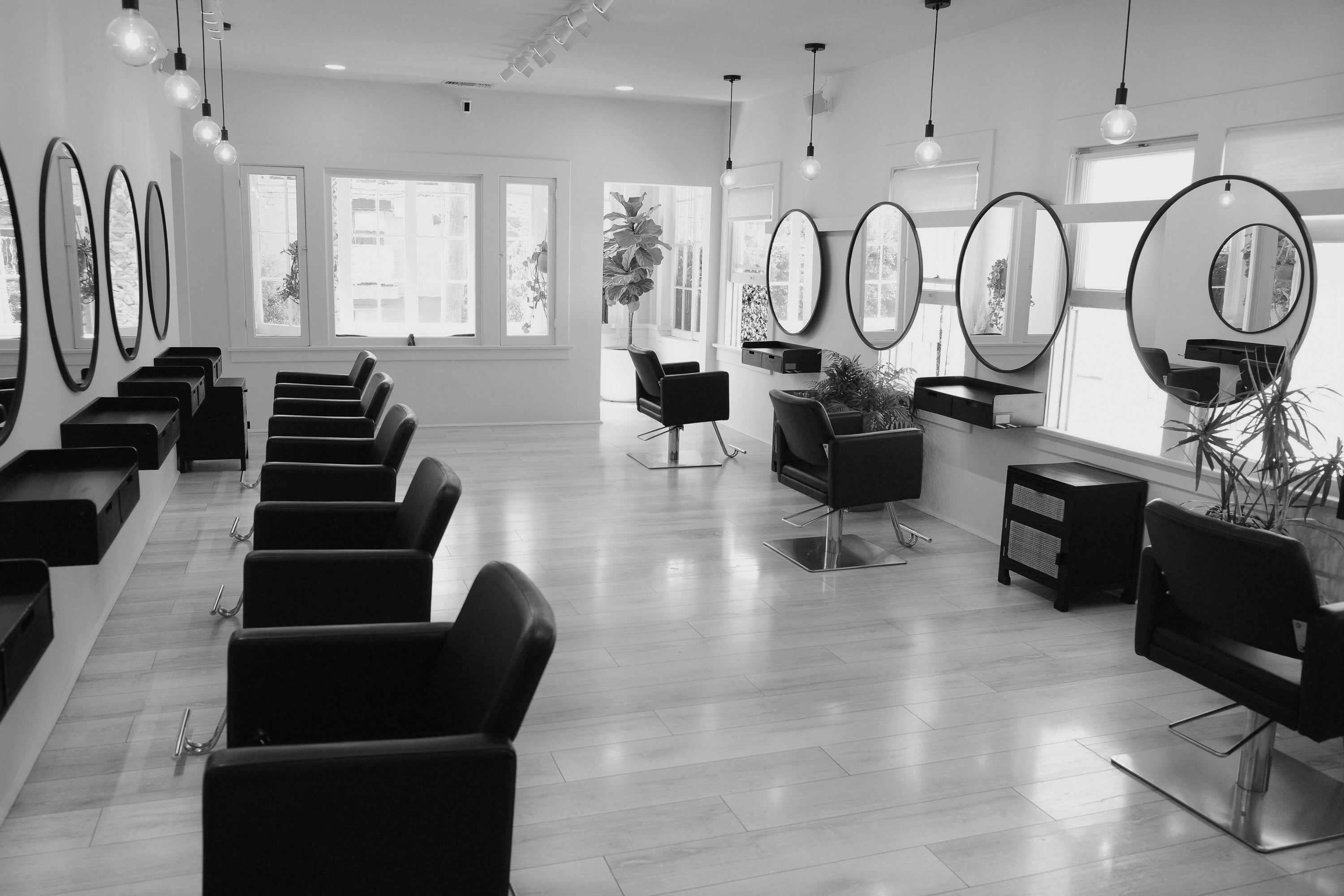 Black and white interior of a modern hair salon with multiple styling stations, black chairs, and round mirrors.