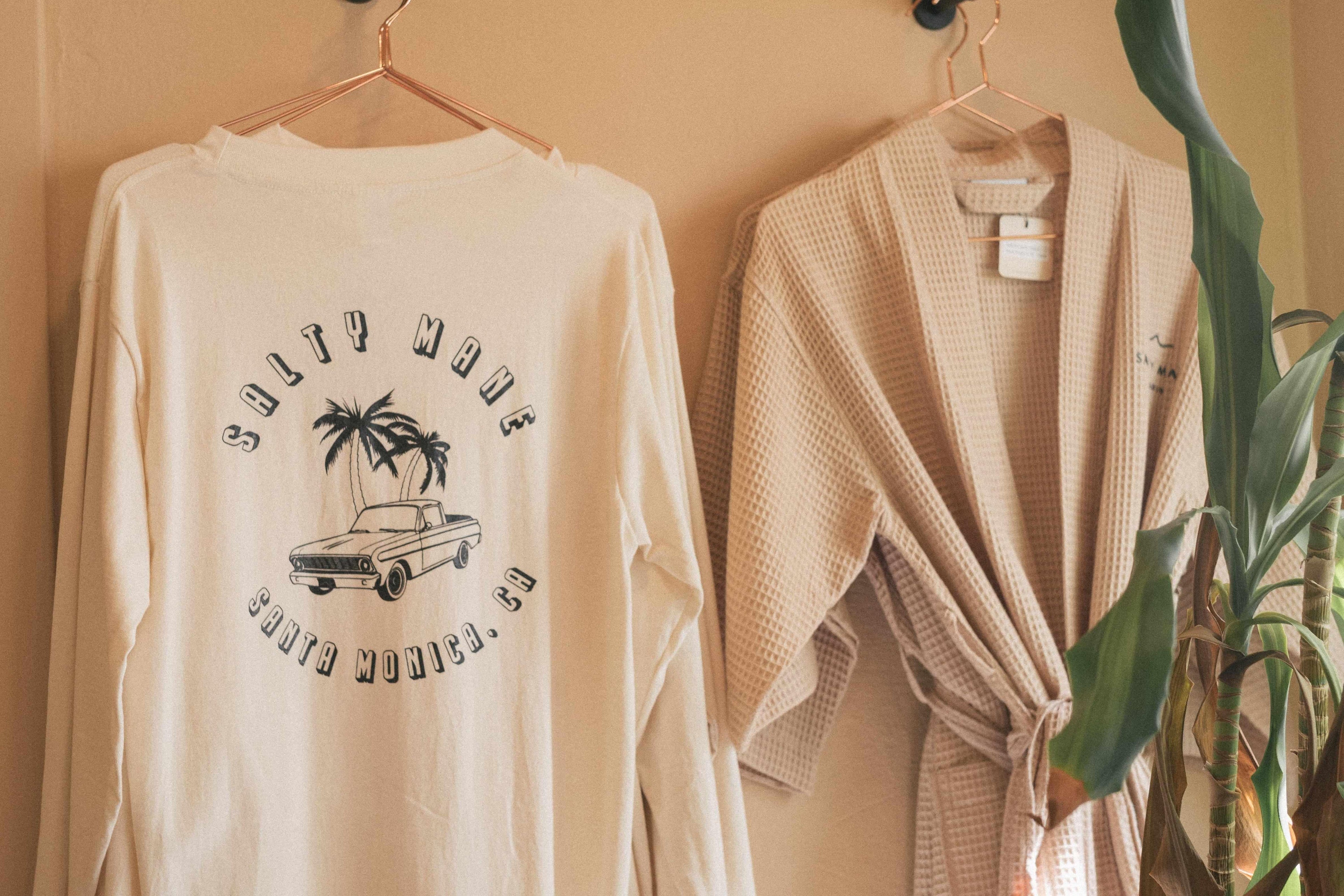 Salty Mane beige long-sleeve shirt with graphic design and beige robe hanging on a hook against a beige wall.