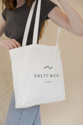 White canvas Salty Mane Studio tote bag with black logo, held by a person in a gray top.