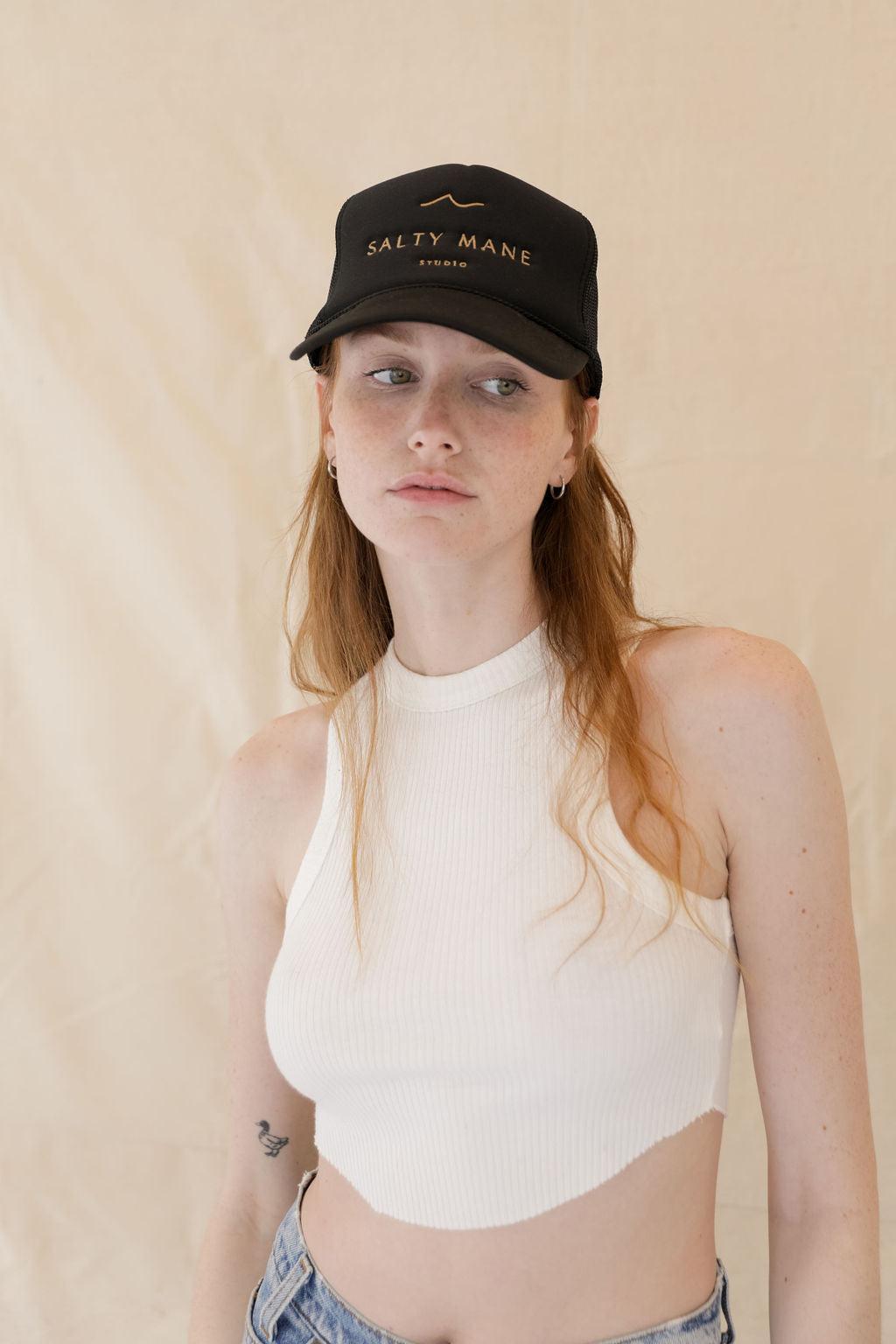Black mesh-back trucker hat with "Salty Mane Studio" in gold text, worn by a person with red hair.