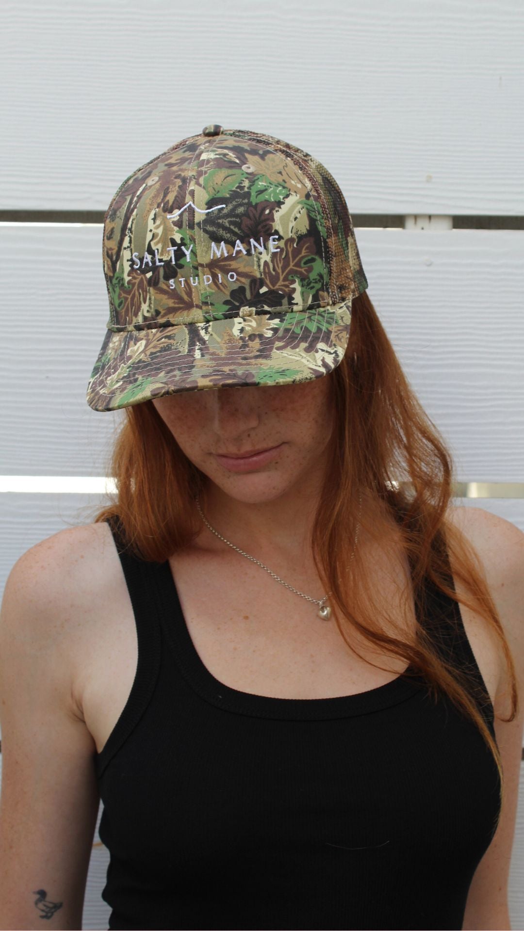 Salty Mane Studio camo trucker hat with mesh back, worn by a person with red hair.