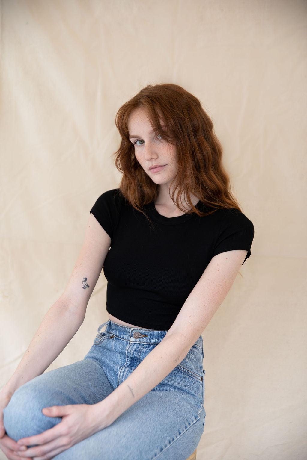 Woman with red hair wearing a black ribbed cropped top and light wash jeans.