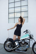 Woman in a black checkered silk mini dress with spaghetti straps, posing on a vintage motorcycle.