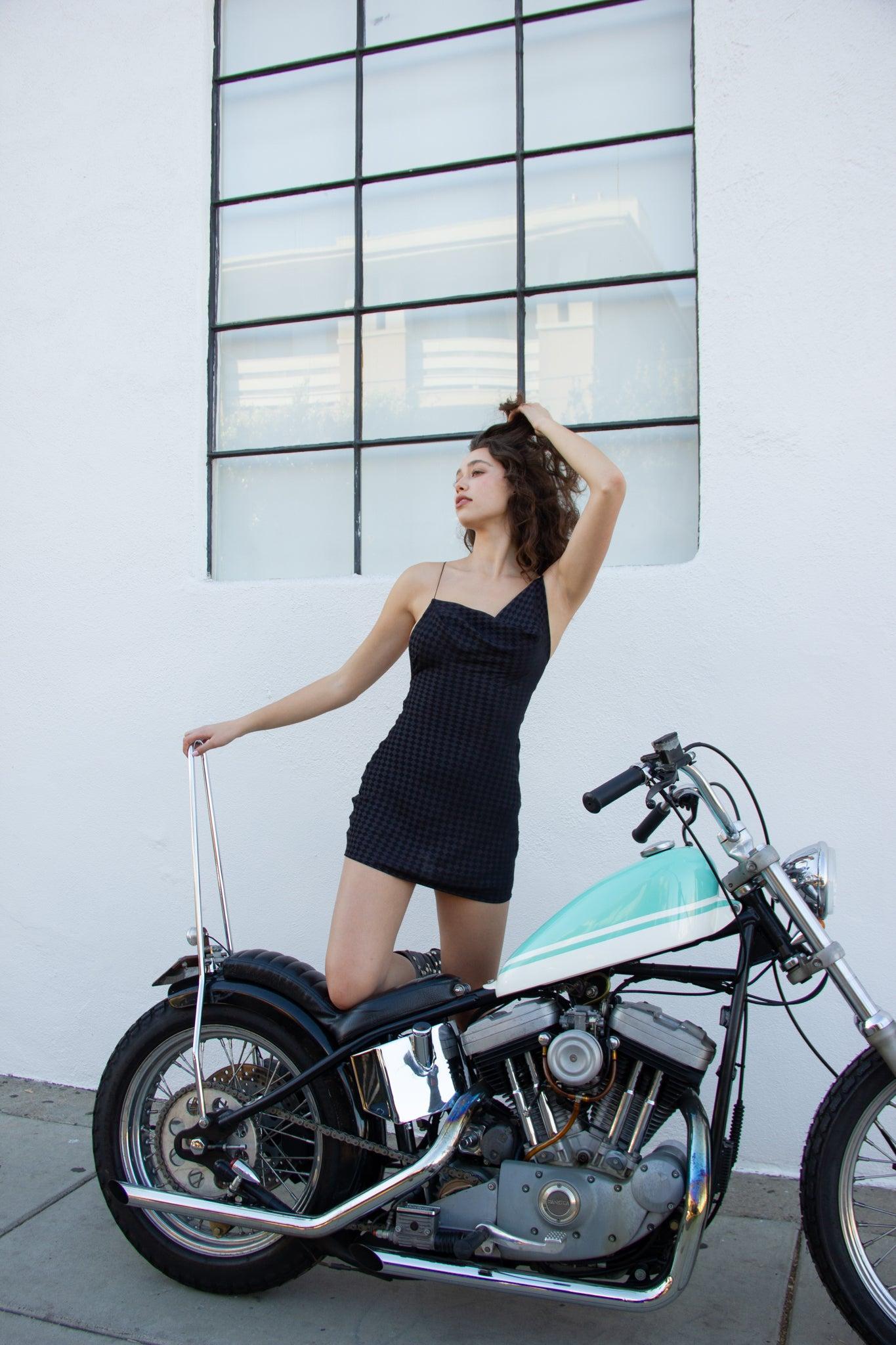 Woman in a black checkered silk mini dress with spaghetti straps, posing on a vintage motorcycle.